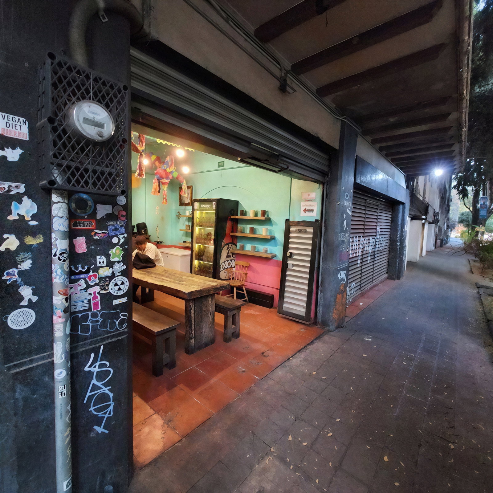 A person sits inside a small, colorful store with vibrant walls, a wooden table, and shelves. The entrance has stickers and graffiti.