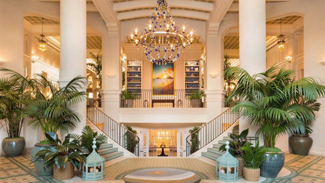 Elegant hotel lobby with grand staircase, lush green plants, blue chandelier, and serene painting. Warm lighting creates a welcoming atmosphere.