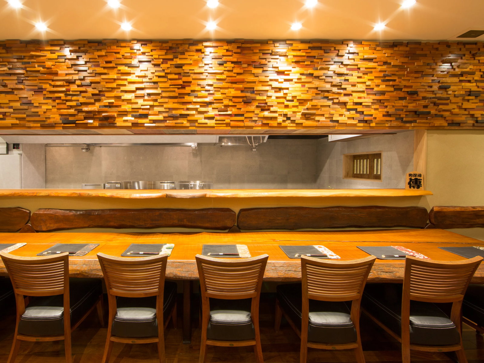 Restaurant interior with a wooden bar and chairs, warm lighting, textured wood wall, and place settings on the counter; cozy ambiance.