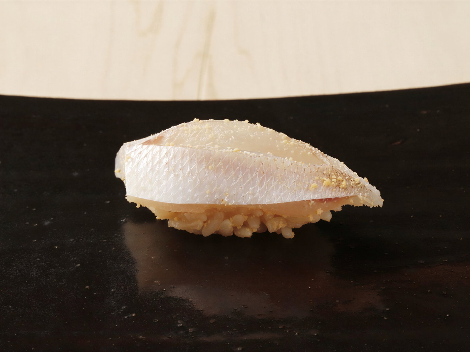 Sushi with a white fish slice on rice, on a dark surface. The fish has a textured, shiny appearance. Simple and elegant presentation.