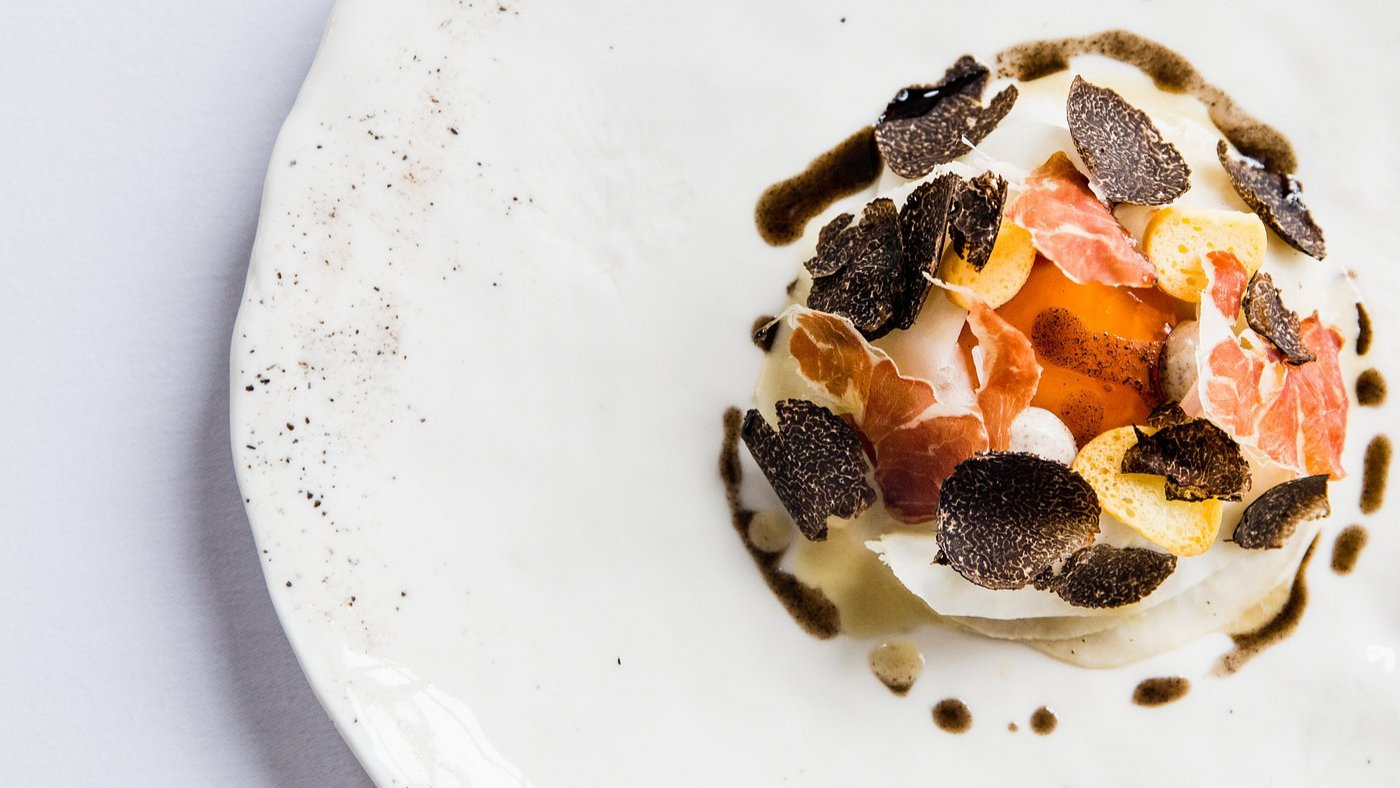 Gourmet dish with an egg yolk, truffle slices, and prosciutto on a white plate, accented by dark sauce drops, creating a refined look.