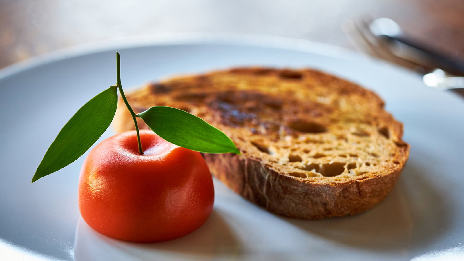 A white plate with a tomato and green leaves, alongside a slice of toasted bread. Fork and knife in the blurred background. Elegant setting.
