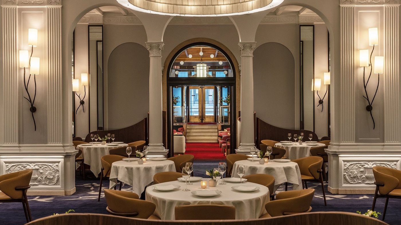 Elegant restaurant with white tablecloths, warm lighting, and decorative columns. Empty tables with wine glasses create a sophisticated ambiance.