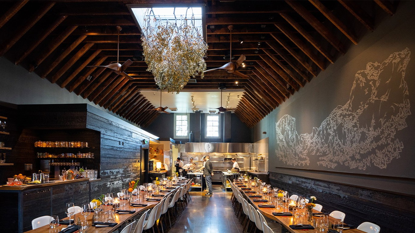 Dimly lit restaurant with a high wooden ceiling and hanging foliage. Long tables set for dining, staff preparing in an open kitchen.