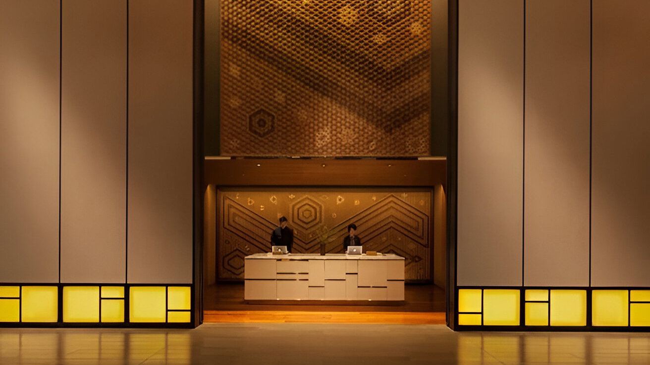 Two people stand behind a modern reception desk in a large, warmly lit lobby with geometric wall patterns and soft yellow accents.