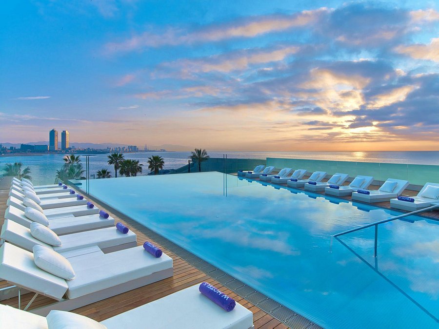 Infinity pool with white loungers and towels overlooking a serene ocean at sunset. The sky is vibrant with clouds, and city buildings are visible.