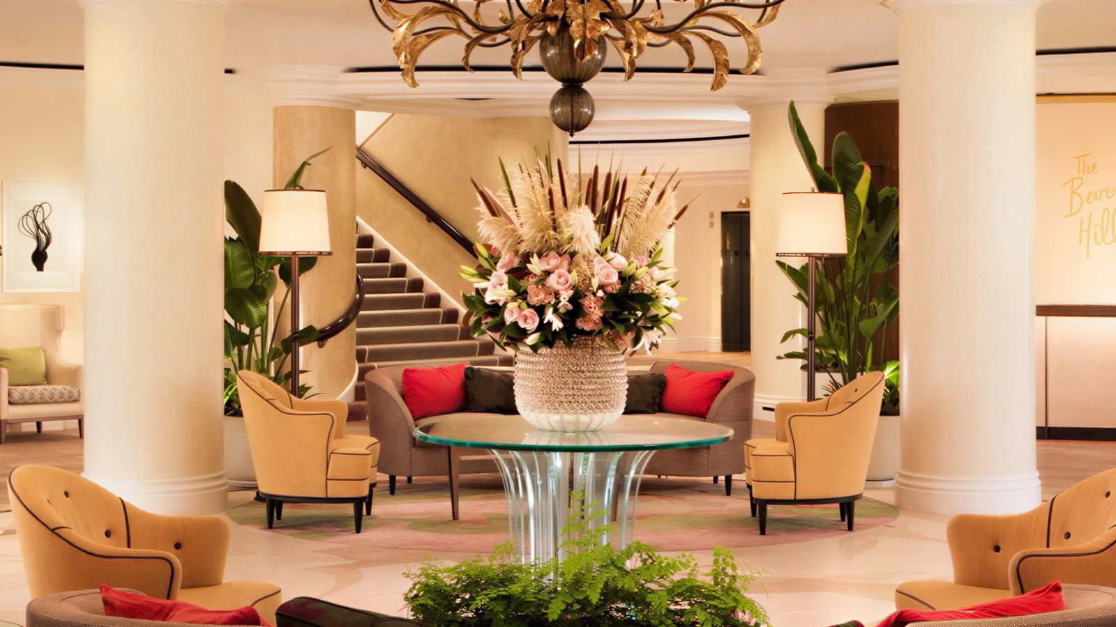 Luxurious hotel lobby with beige chairs, a glass table, and a large floral arrangement. Warm lighting, green plants, and stairs in the background. (AI-generated, newly added)