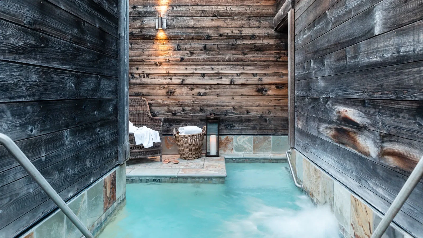 Cozy spa setting with a steaming turquoise pool lined by wooden walls. A wicker chair, basket, and towel evoke relaxation. Warm lighting.