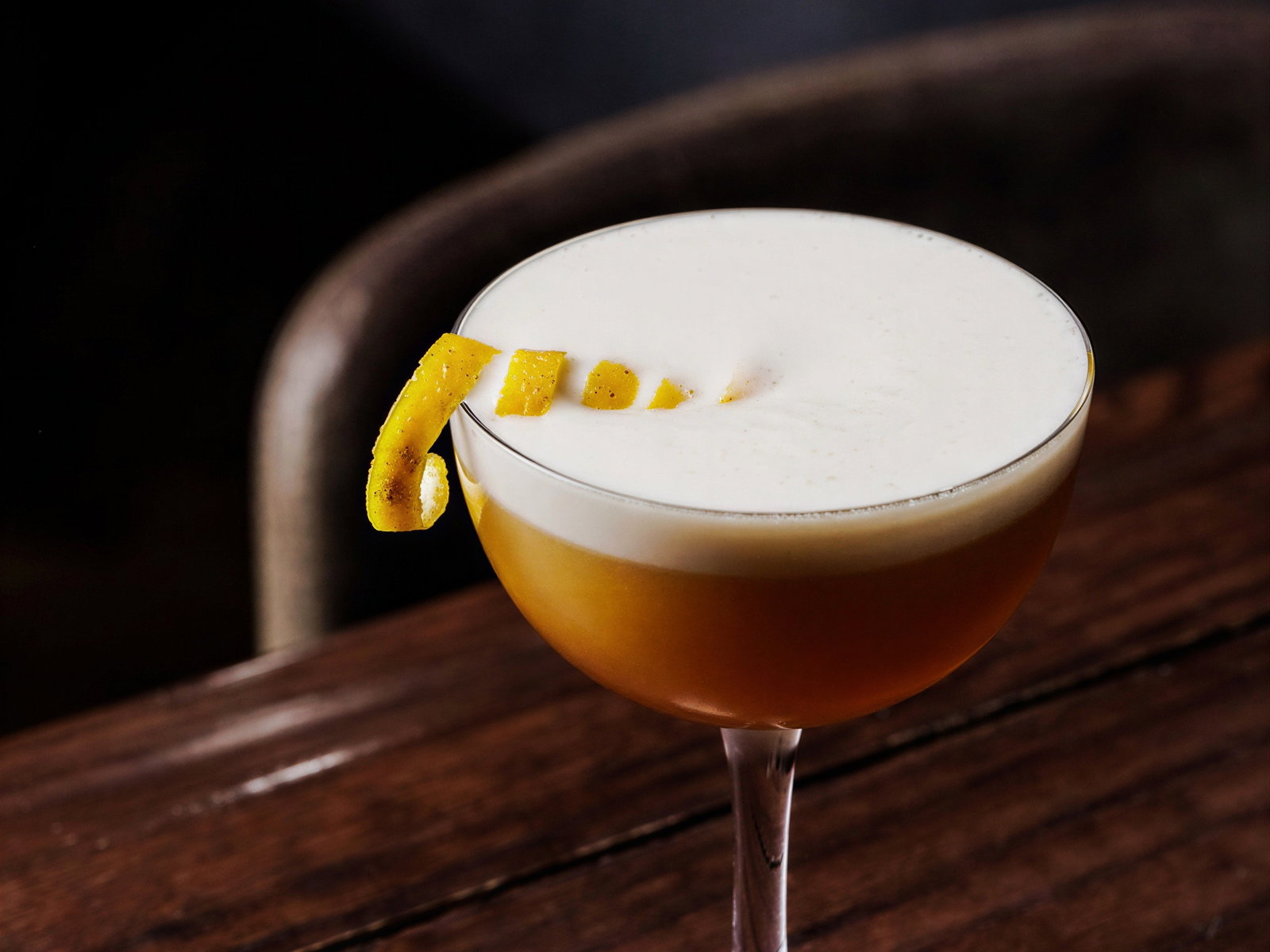 A cocktail with a frothy top and lemon twist garnish on a wooden table, set against a blurred dark background.