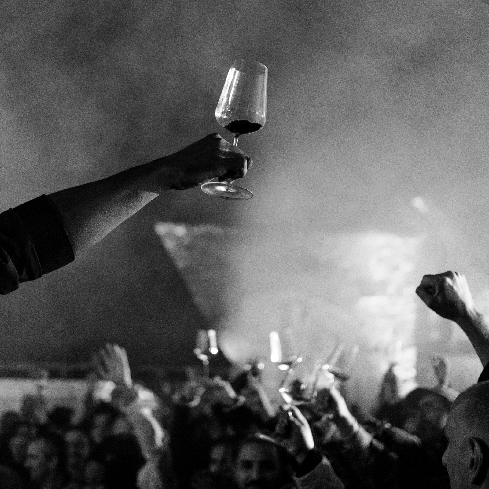Vin et hip hop (wine and hip hop) Crowd celebrates at a smoky event, raising wine glasses high. B&W setting, festive atmosphere with outstretched arms and cheers.