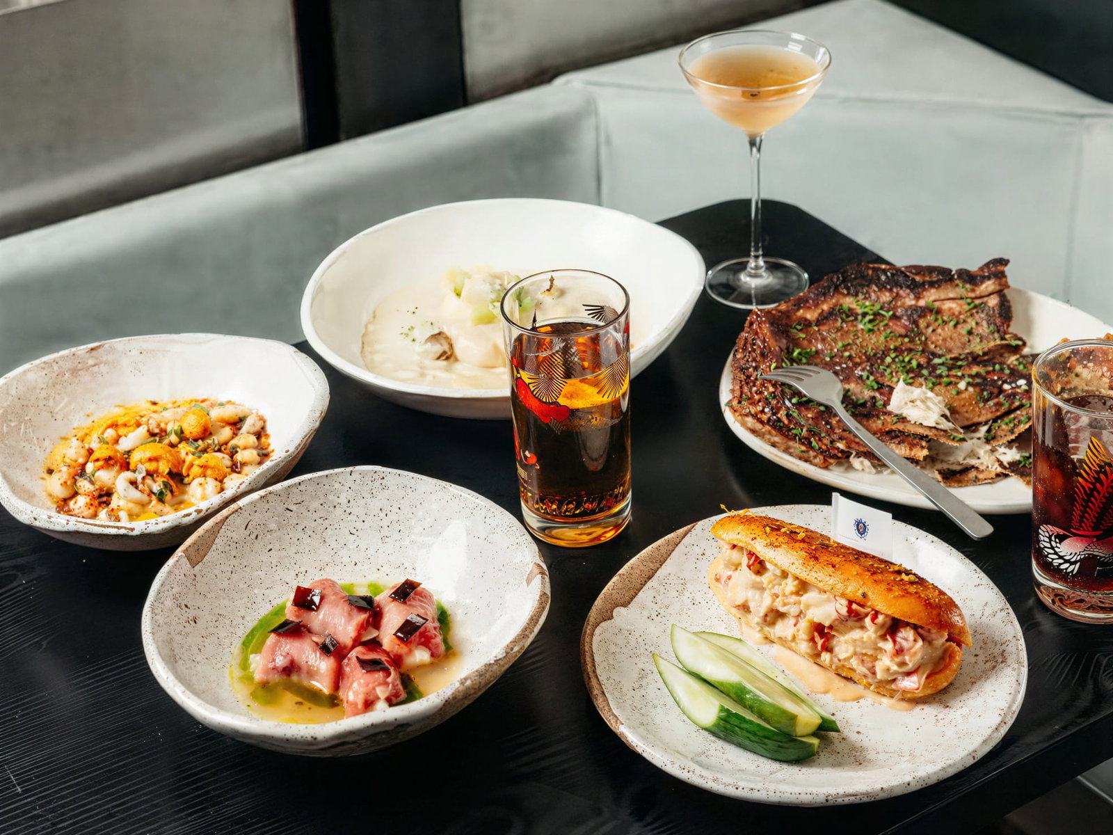 A table with a selection of dishes from Smithereens NYC including seafood, meat, and cocktails. Background has a gray sofa. Plates are speckled, and mood is elegant.