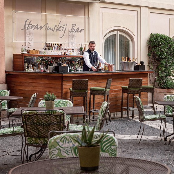 Bartender prepares drinks at an outdoor bar with green, leafy-patterned chairs. "Stravinskij Bar" sign above; cozy, elegant setting.