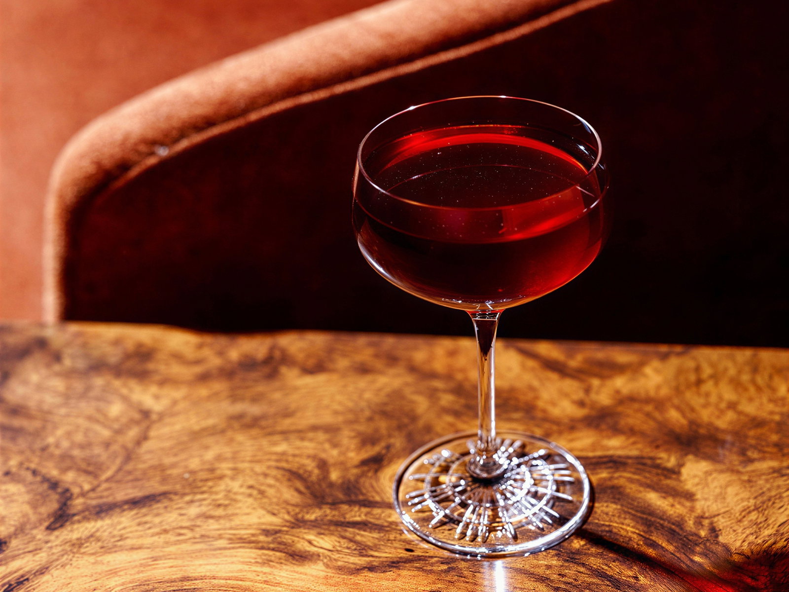 A red cocktail in a crystal glass on a wooden table, with a plush brown chair in the background, evokes a warm, elegant setting.