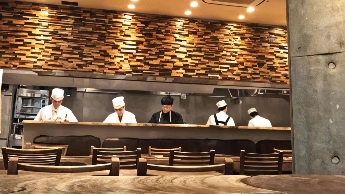 Chefs in white uniforms work at a wooden counter in a modern restaurant. The warm, textured wall and wooden furniture create a cozy atmosphere.