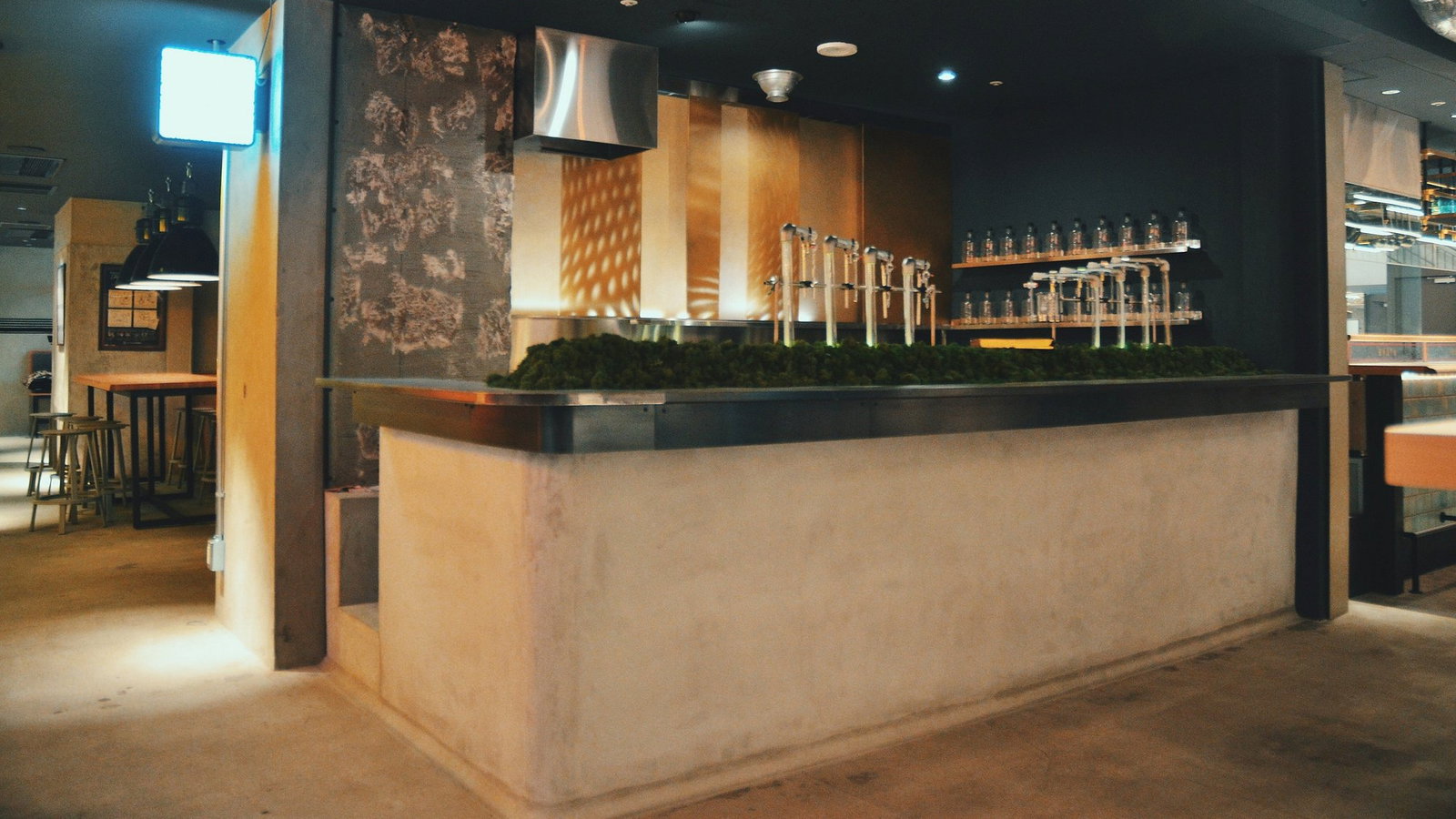 Modern bar interior with concrete counter, metal taps, and bottles on shelves. A blue-lit sign, industrial lights, and wooden tables. Dimly lit.