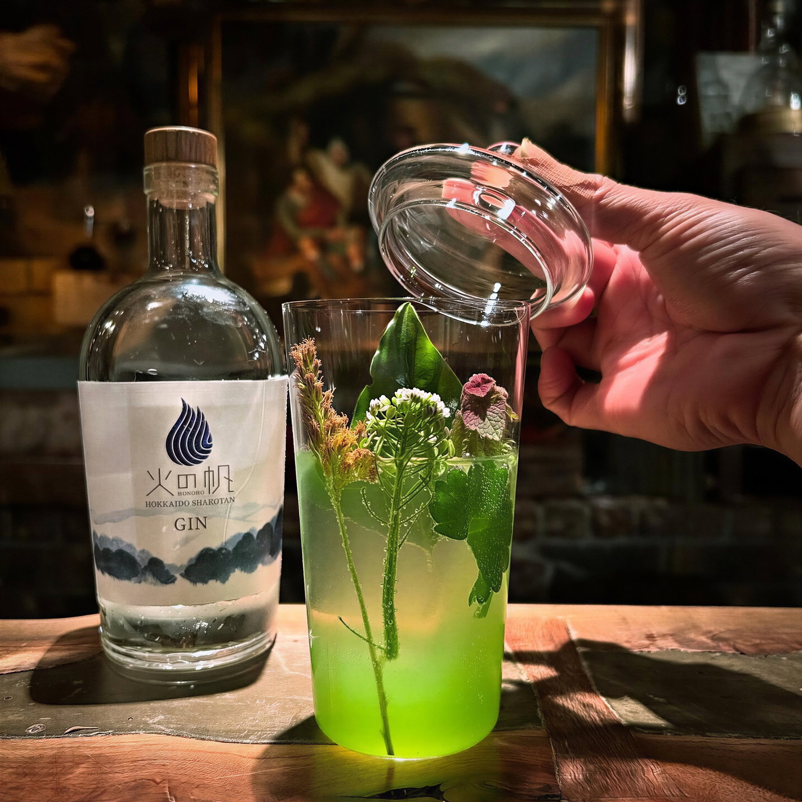 Hand holding lid above glass with green drink, herbs, flowers; bottle of gin labeled "HOKKAIDO SHIROTAN" nearby. Warm, rustic setting.