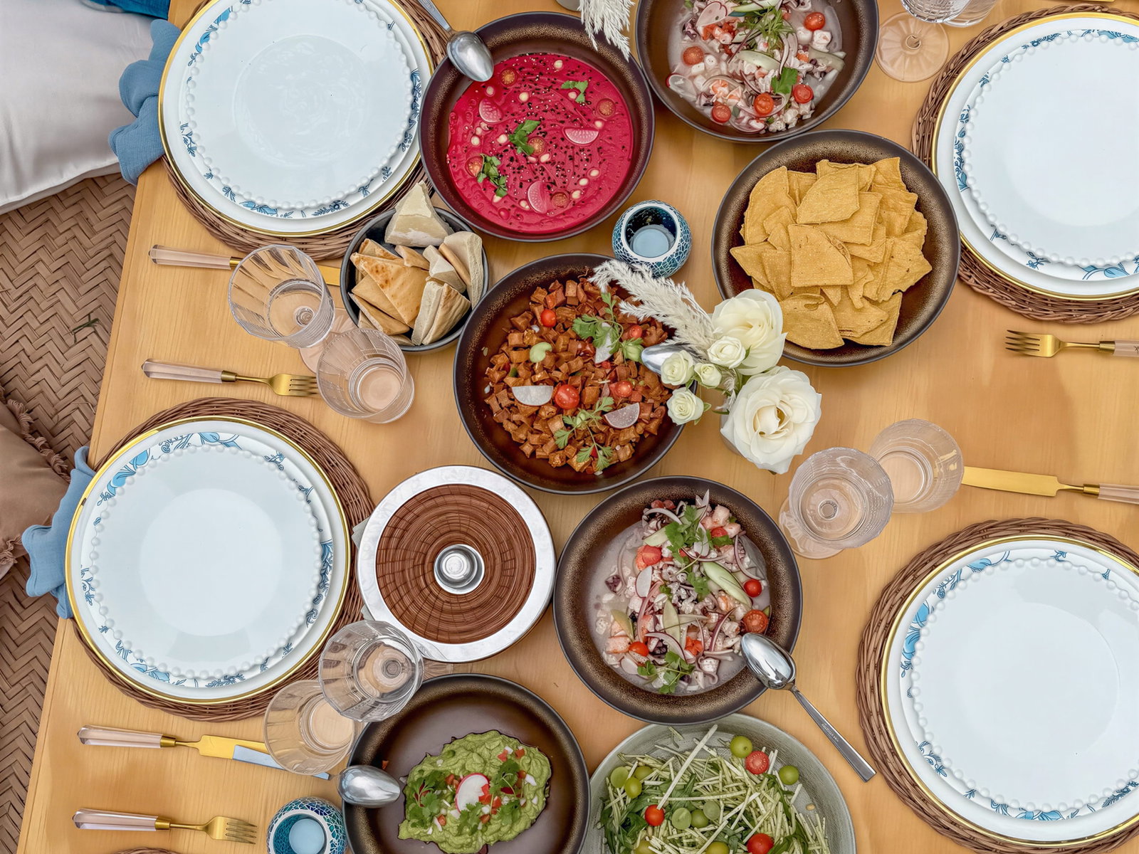 A wooden table with assorted dishes from El Cocal at Wakax Hacienda including beet soup, chips, salads, and flowers. Six place settings with blue-patterned plates.