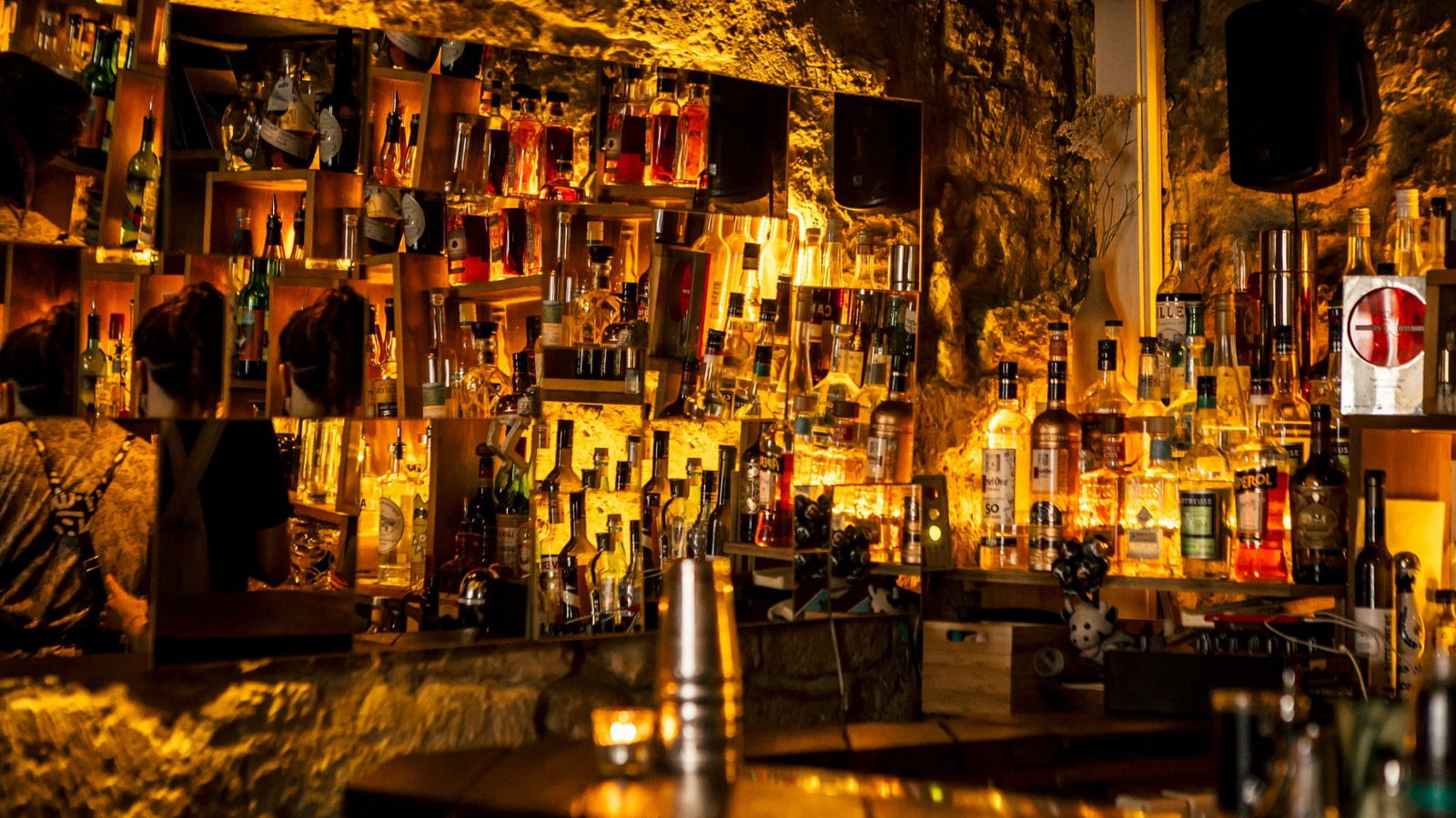Dimly lit bar with mirrored shelves reflecting various liquor bottles, creating a warm, cozy ambiance. A person stands at the counter.