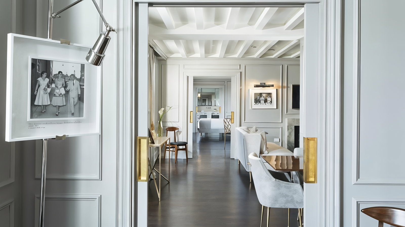 Elegant room with white and gray decor, featuring a black-and-white photo, modern furniture, and a view through open doors to a bed.