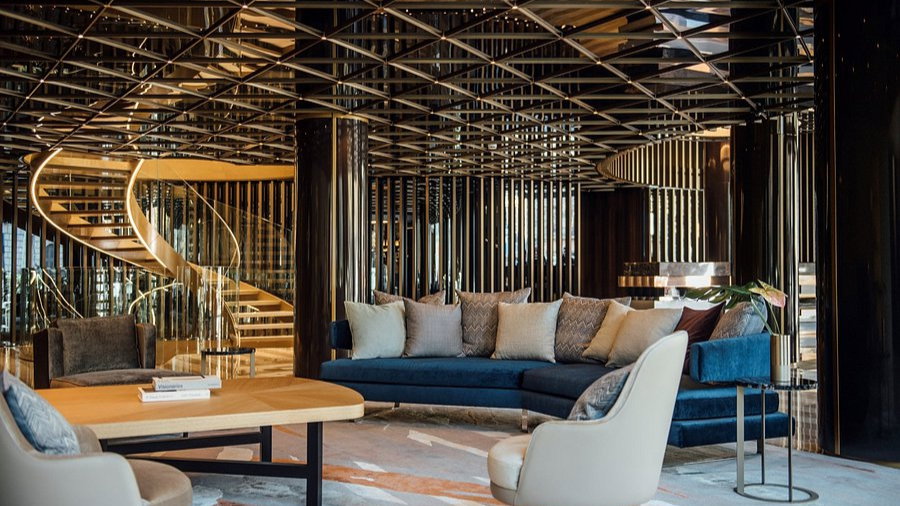 Luxurious lounge with blue sofas, beige chairs, and a wooden table. Elegant spiral staircase and reflective ceiling create a modern feel.