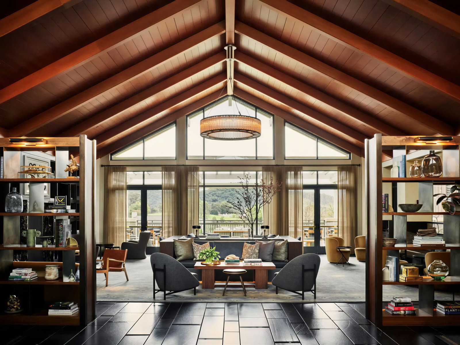 Spacious room with vaulted wood ceiling, large windows, and bookshelves. Modern furniture in neutral tones. Scenic view outside. Cozy ambiance.