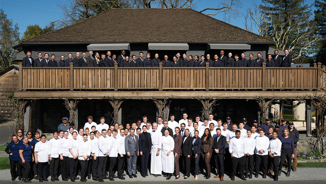 A large group of people in chefs outfits and suits stand in front of an older brown building.