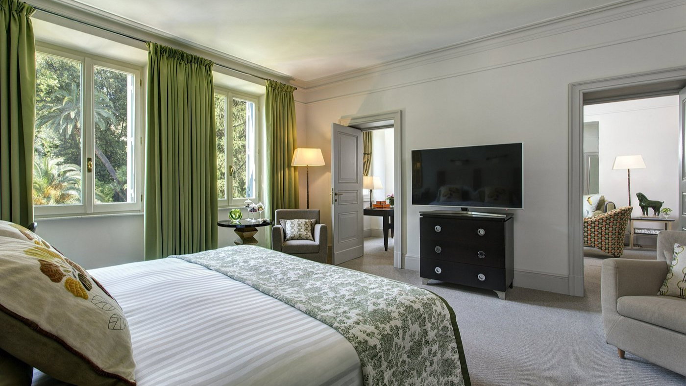 Elegant hotel room with a bed, green curtains, and a TV. Sunlit windows overlook trees. Cozy seating area with a lamp, serene atmosphere.