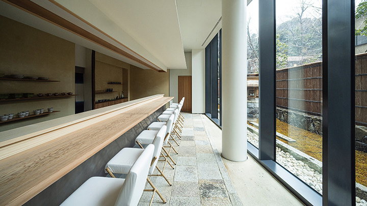 Minimalist dining area with a long wooden bar, white chairs, and large windows. Stone flooring and natural light create a serene atmosphere.