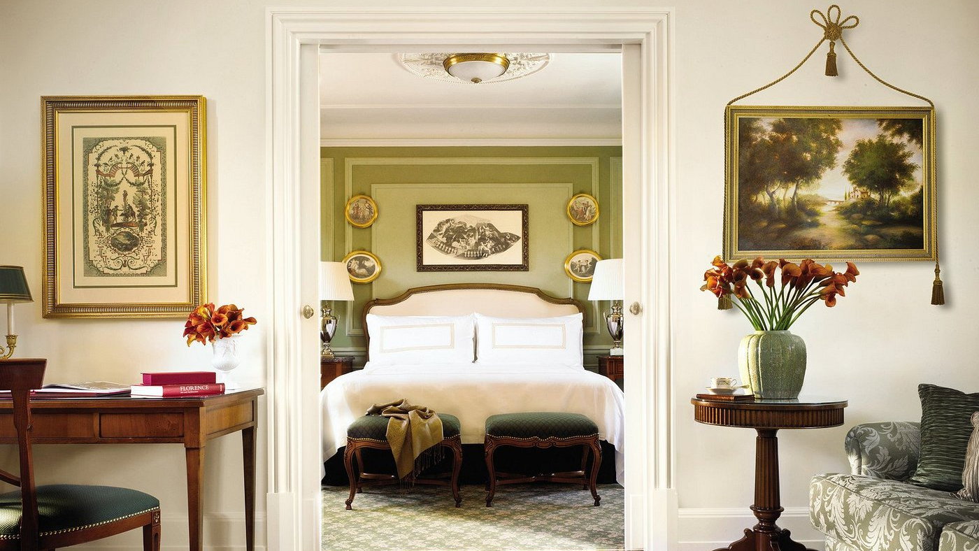 Elegant bedroom with a large bed, green walls, floral artworks, and vintage decor. Orange flowers and books on a wooden table. Cozy ambiance.