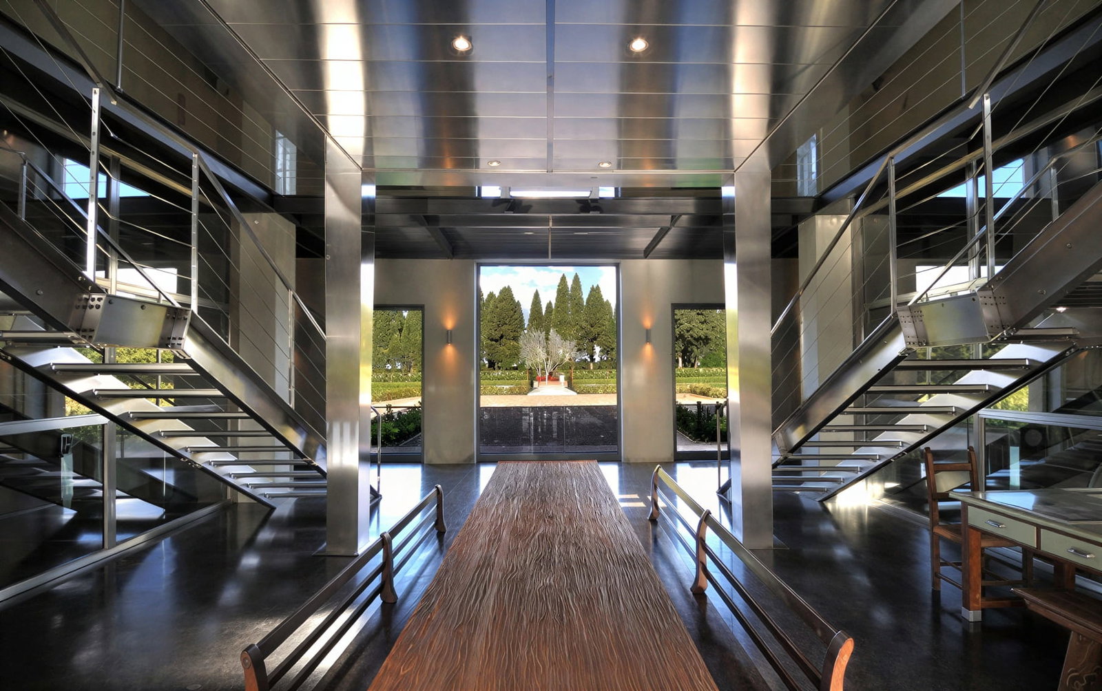 Modern interior entrance of Tenuta San Guido with stainless steel stairs, wooden table, and benches. Glass doors open to a garden view. Bright, sleek design.