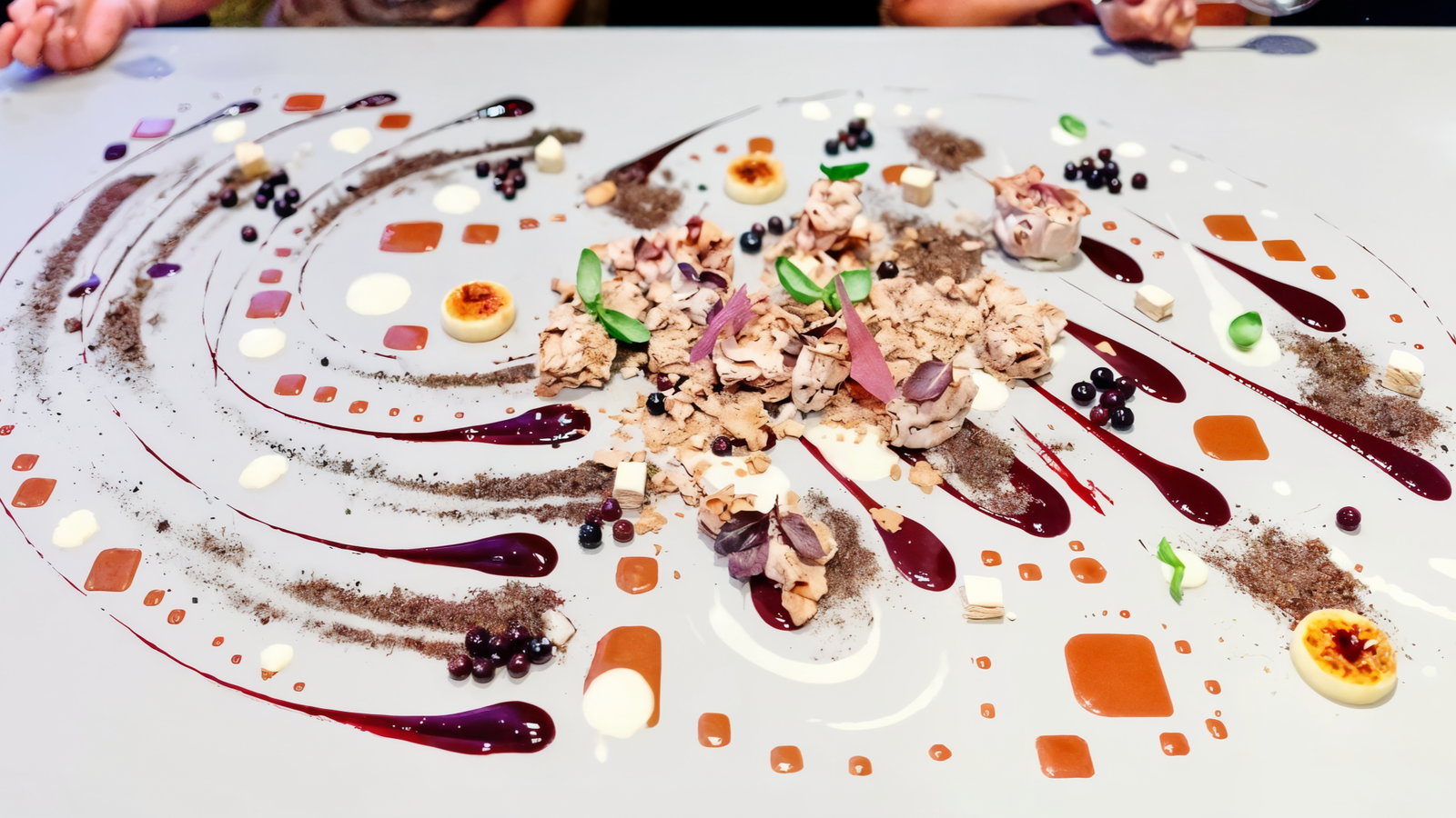 Gourmet dessert artfully spread on a table; various sauces, crumbs, and berries form colorful patterns. Hands visible at the top edge.