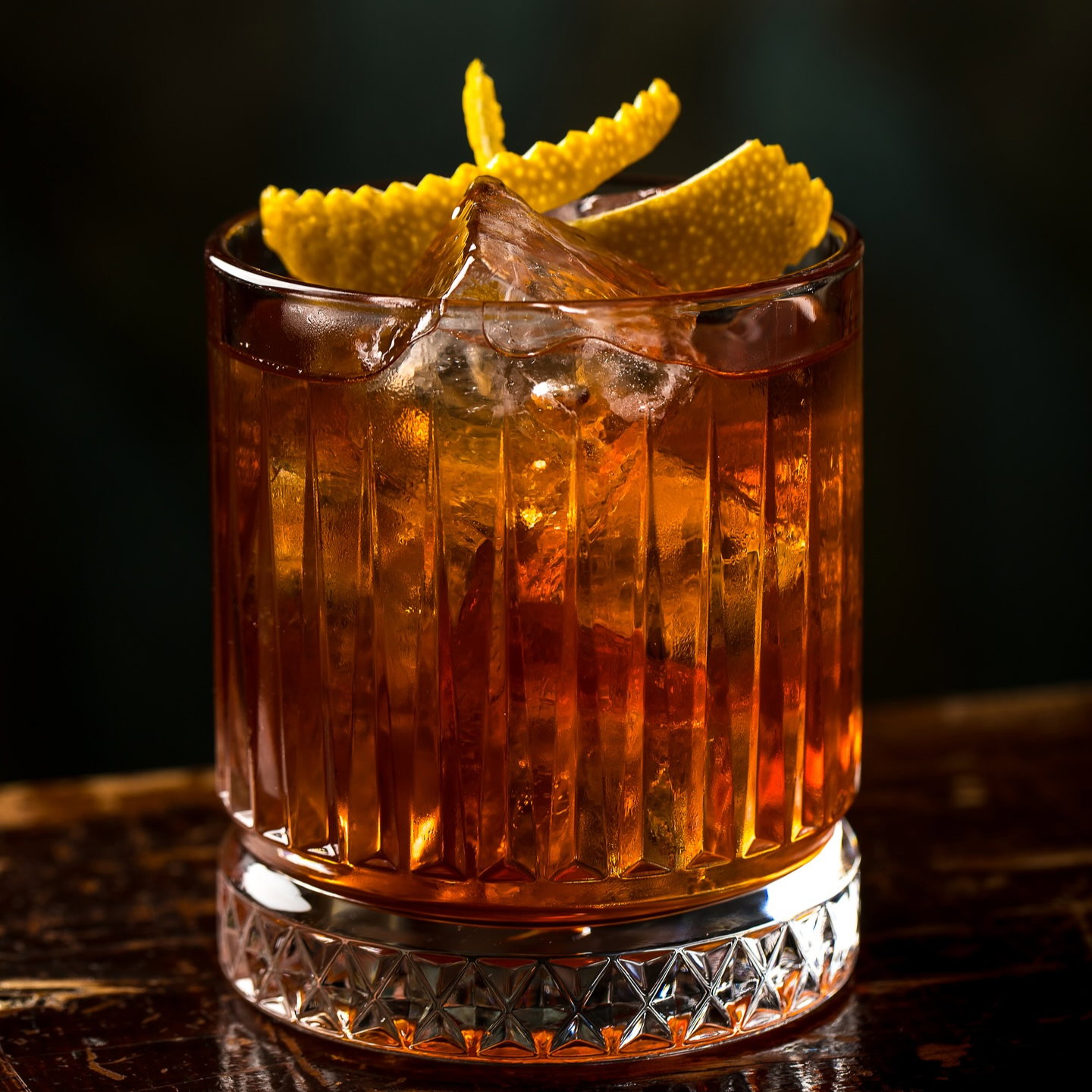 A textured glass of amber liquid with ice and a citrus twist garnish on a wooden table, dark background, elegant presentation.