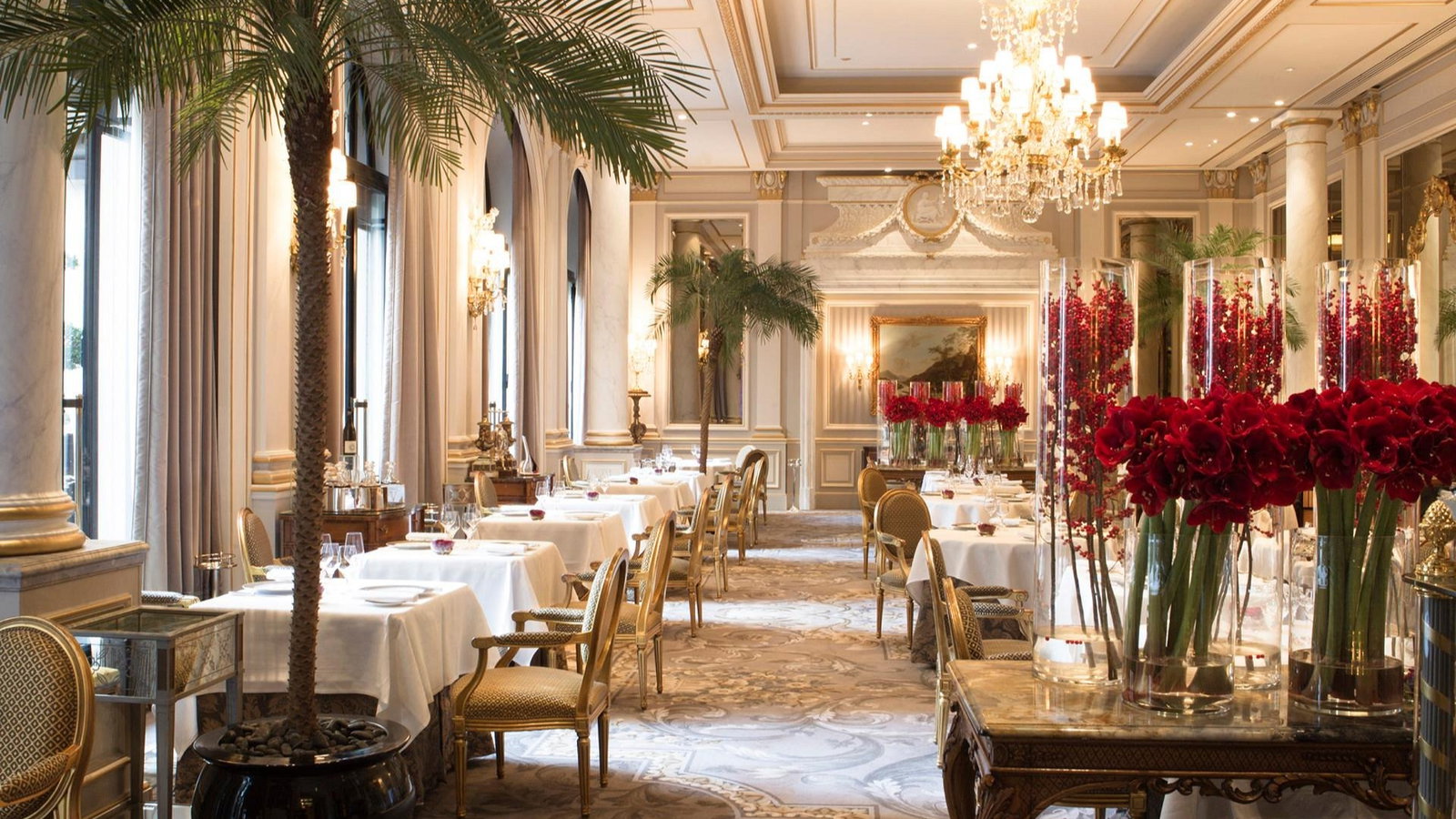 Elegant restaurant interior, featuring white tablecloths, red floral arrangements, chandeliers, and palm trees; a luxurious and serene ambiance.