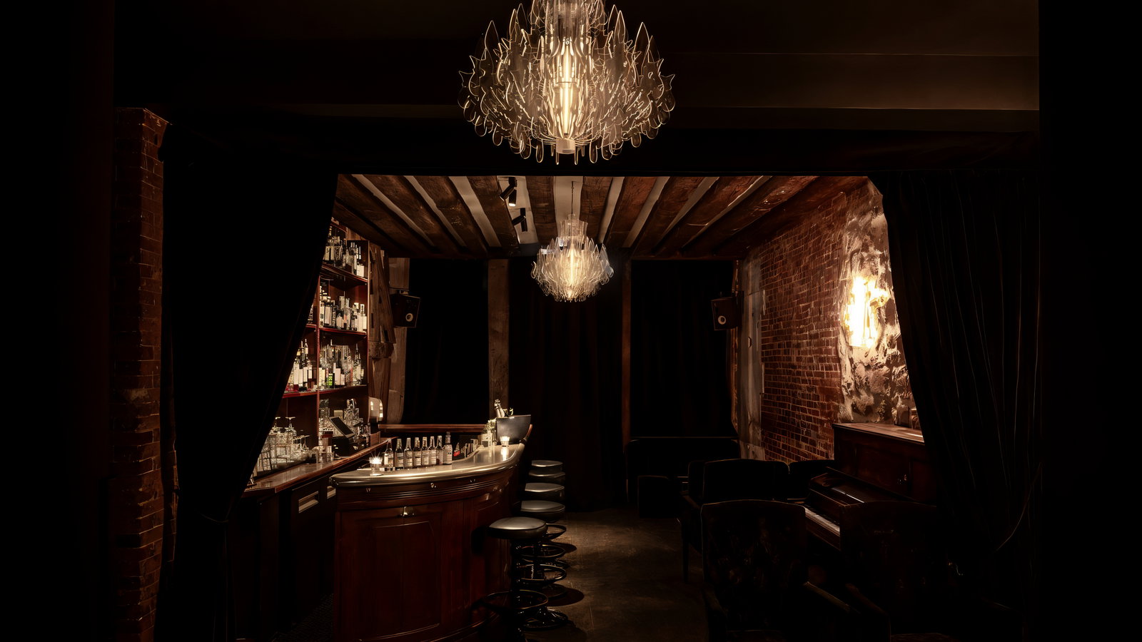 Dimly lit bar with a curved wooden counter, black stools, and shelves full of bottles. Brick walls and a chandelier create a cozy atmosphere.