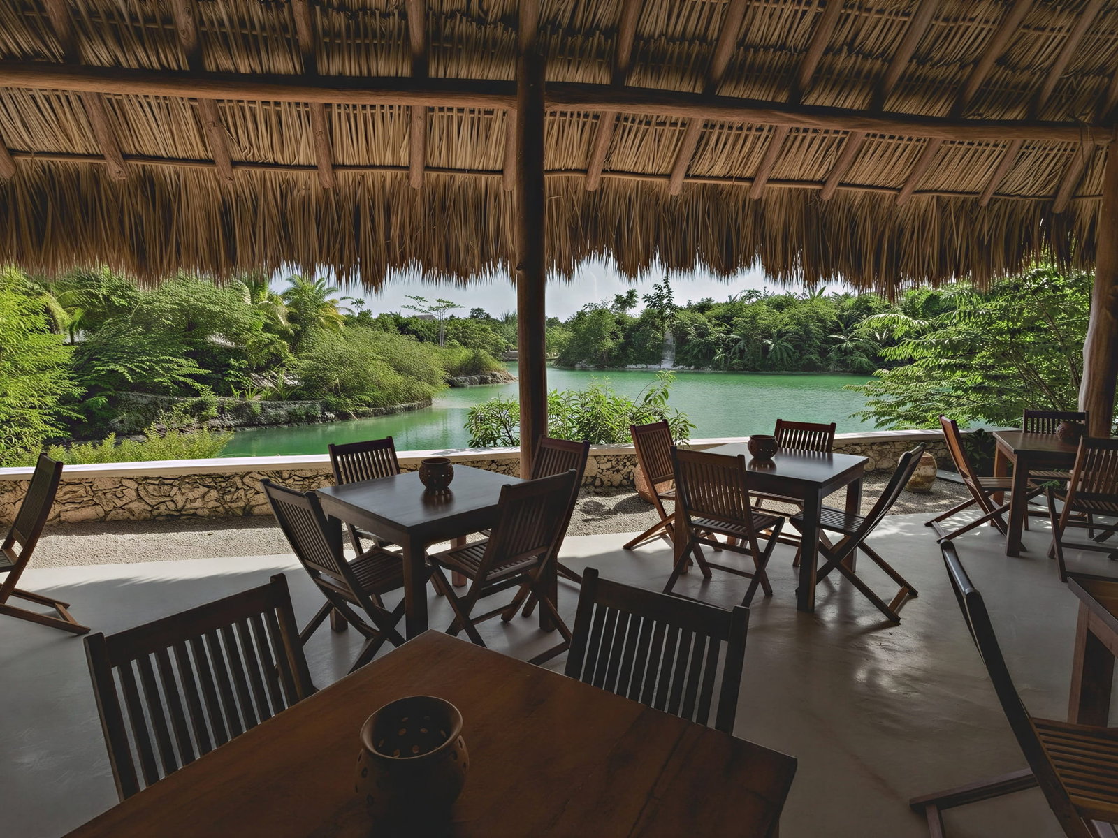 Outdoor seating of El Cocal at Wakax Hacienda under a thatched roof overlooks a serene green lagoon with lush foliage. Empty wooden tables and chairs create a tranquil mood.