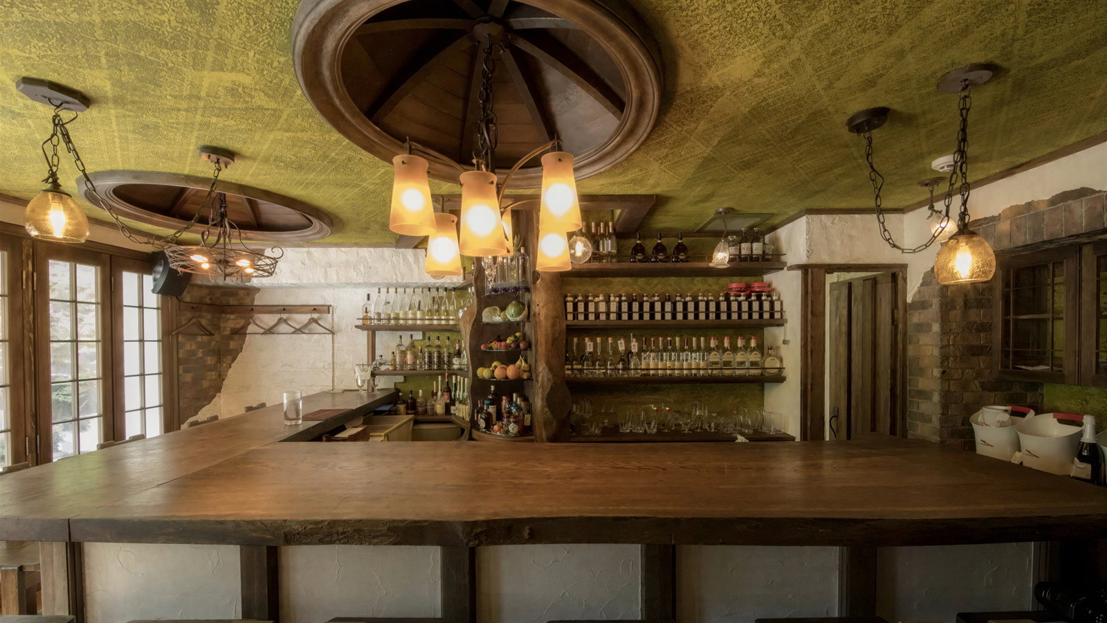 Rustic bar interior with a wooden counter, green ceiling, and warm lighting. Shelves stocked with bottles. Cozy and inviting atmosphere.
