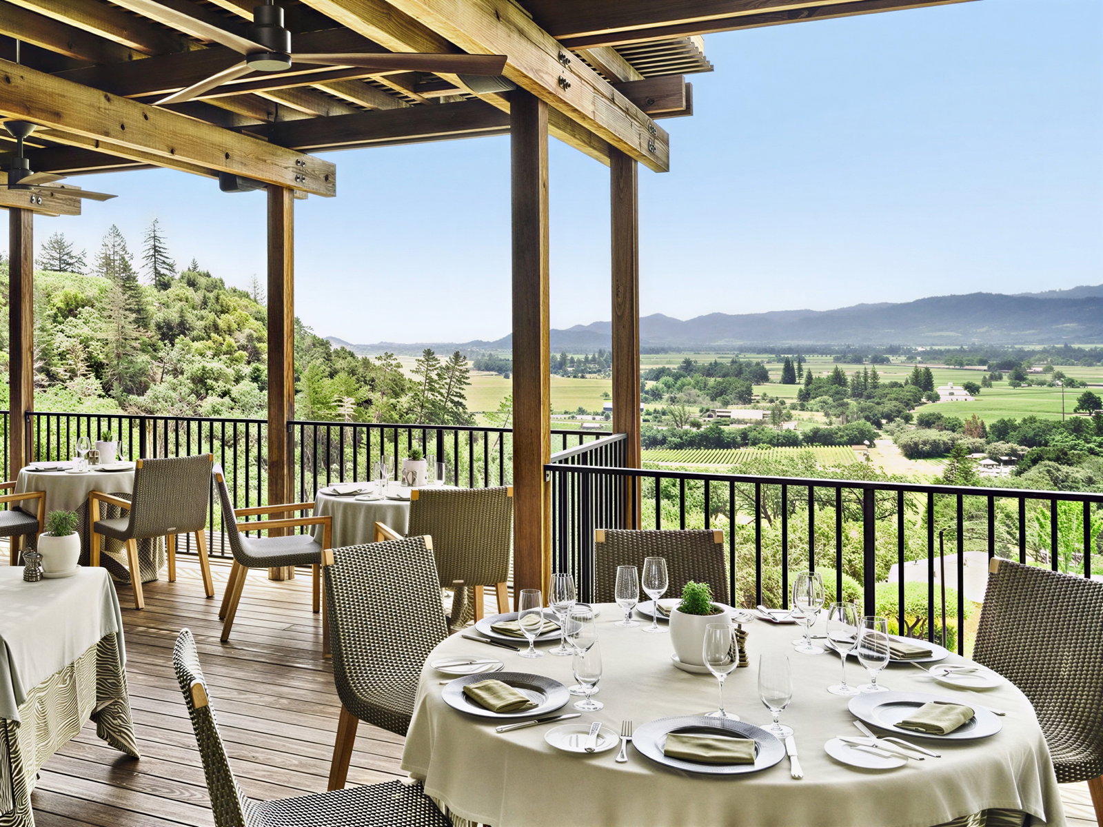 Outdoor restaurant with set tables overlooks lush vineyard and hills. Neutral tones, serene ambiance, clear sky with distant mountains.
