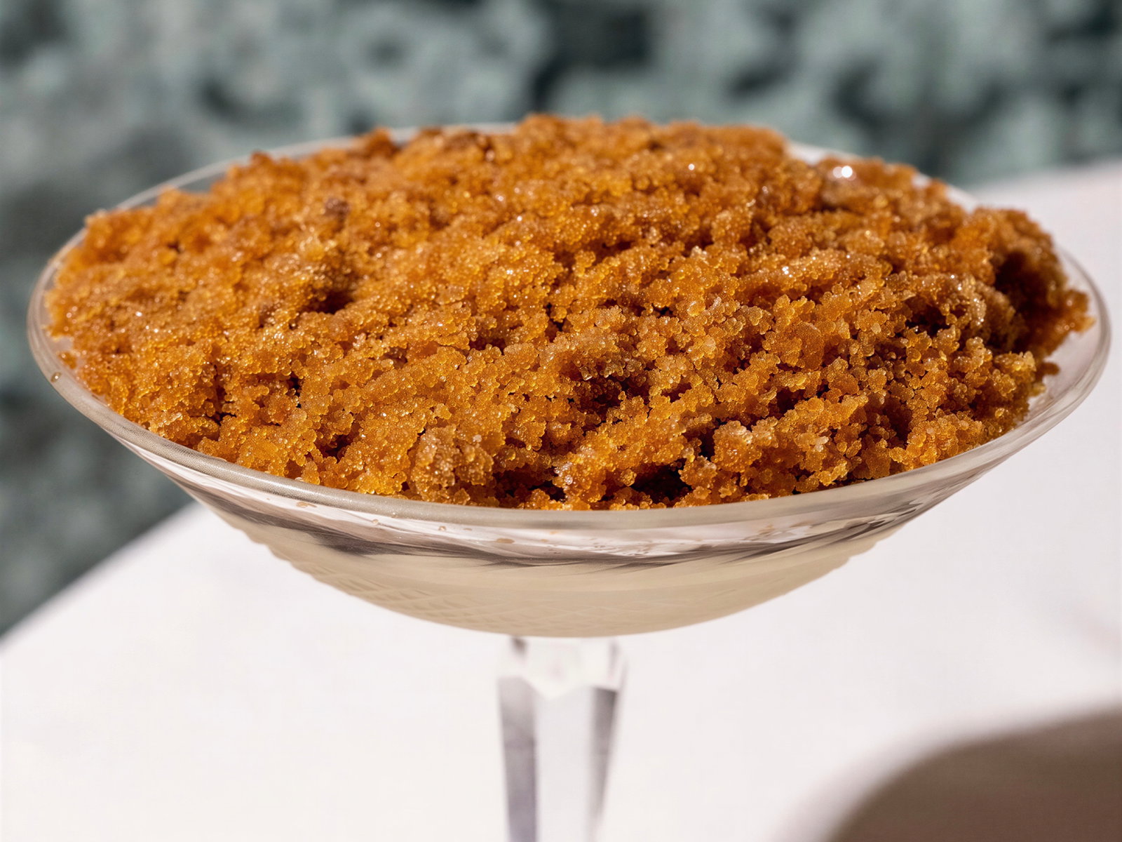 Glass bowl of brown sugar crystals on a white table, with a blurred gray background. Bright lighting highlights the sugar texture.