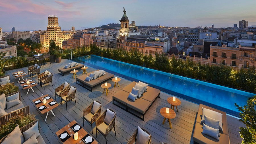 Rooftop terrace with modern seating, dining tables, and a pool overlooking a cityscape at dusk. Soft lighting and a tranquil mood.