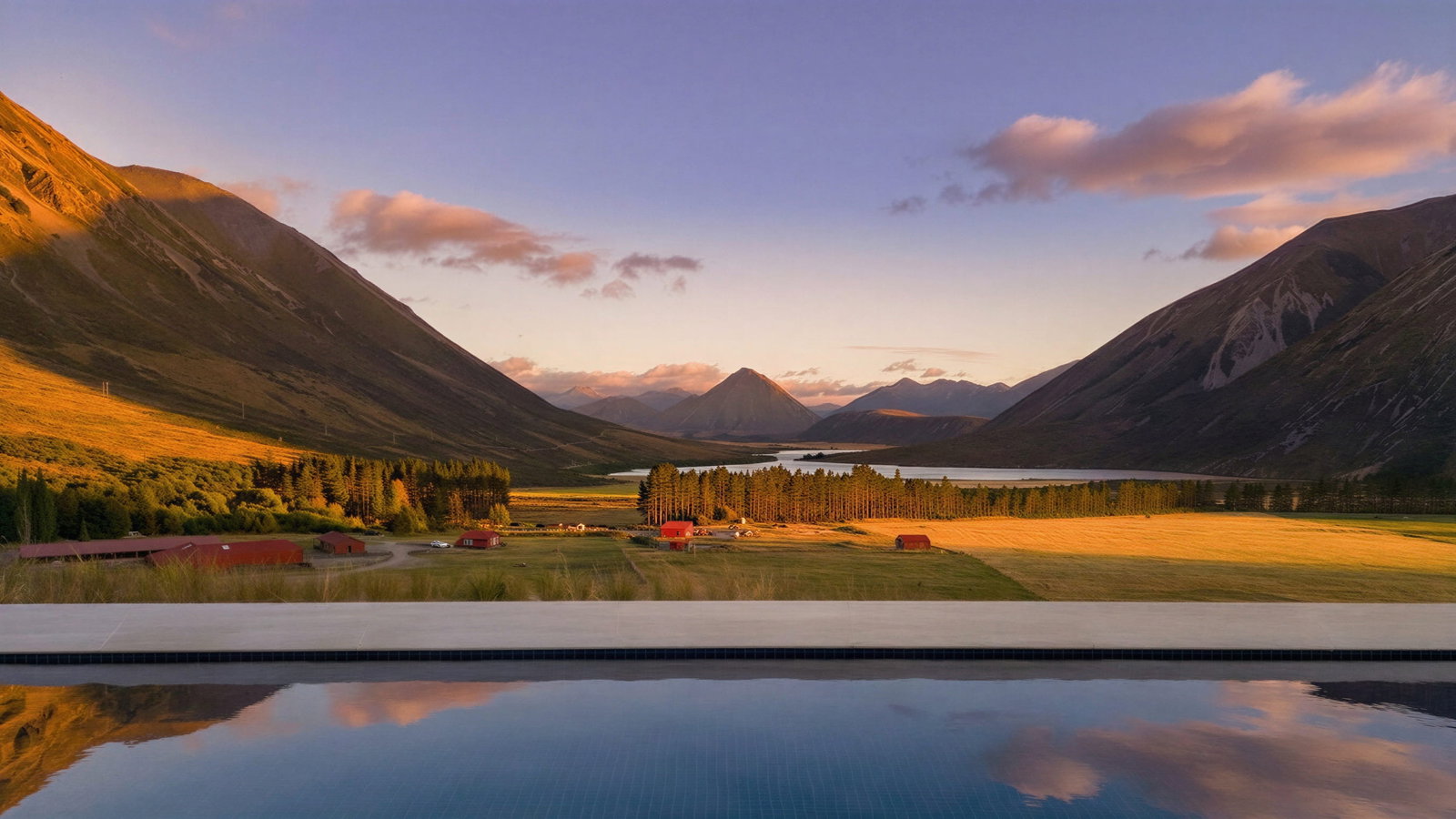 Swimming pool mountain valley view at sunset at Flockhill Lodge, with a calm pool reflecting the sky. Red huts and trees dot the golden fields, creating a peaceful scene.
