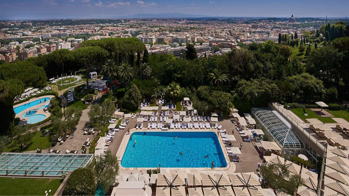 Aerial view of a lively outdoor pool area with sun loungers and people swimming, surrounded by trees and overlooking a vast cityscape.