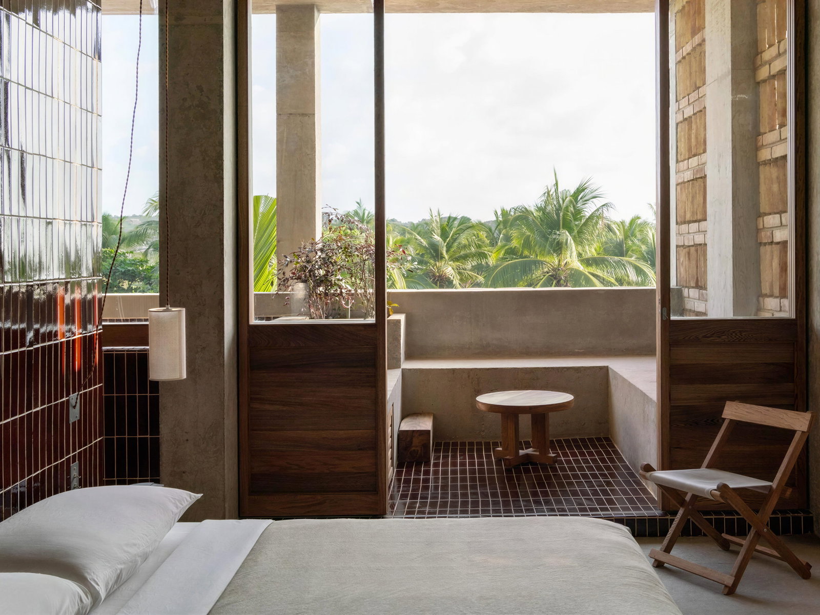 Minimalist bedroom of Ocean View Suite at Hotel Humano with a bed, wooden chair, and round table. Open balcony shows lush palm trees. Neutral tones create a serene vibe.