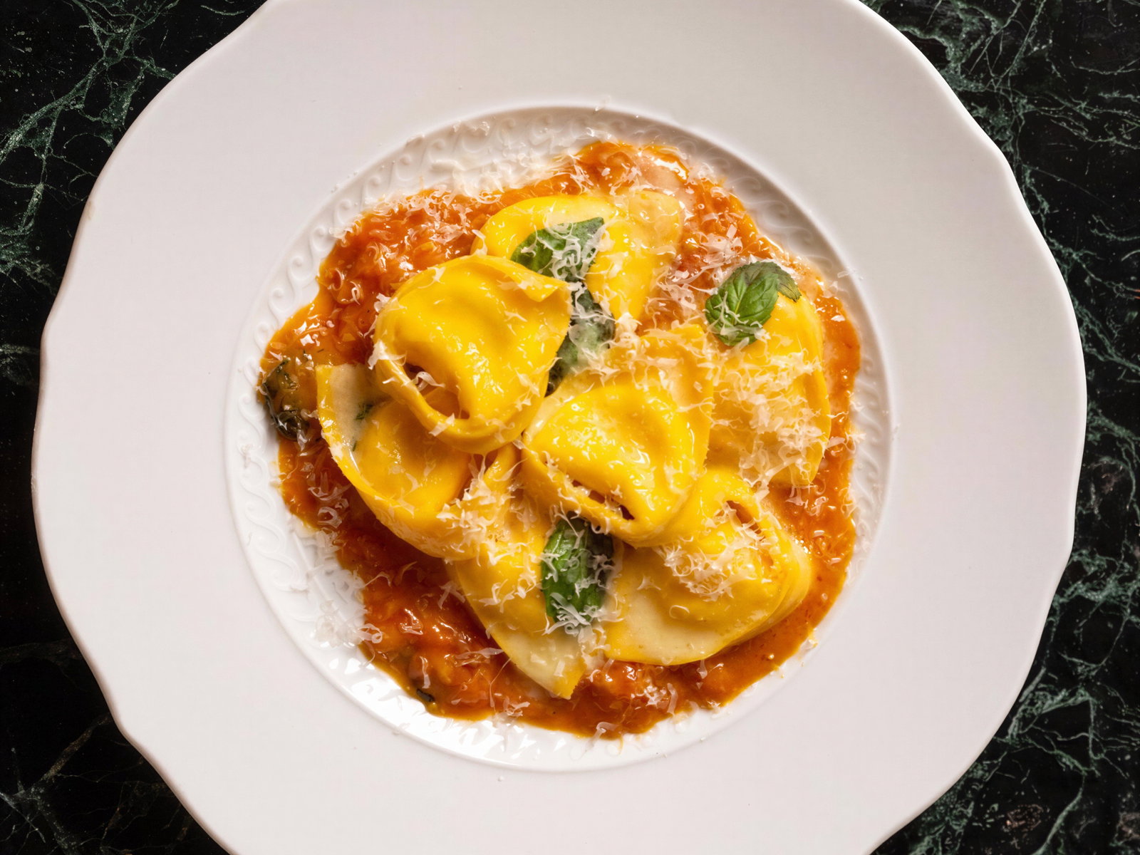 Yellow tortellini on a white plate with vibrant tomato sauce and basil garnish, set on a dark green marble background.