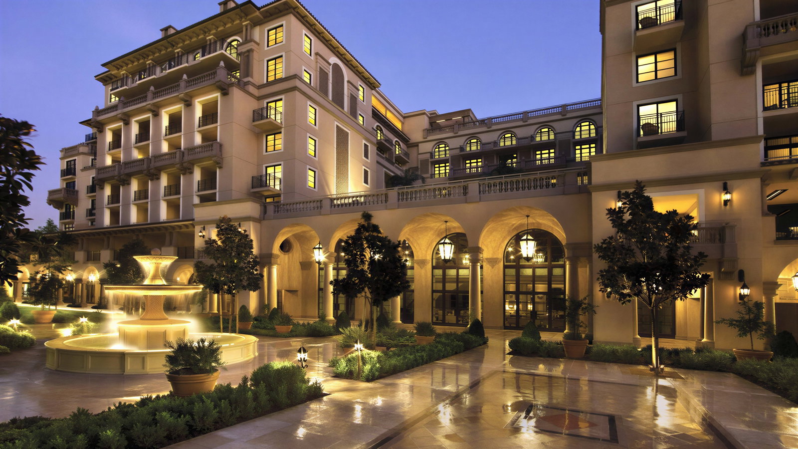 Elegant hotel courtyard at twilight, featuring a lit central fountain, arched entrances, illuminated windows, and lush greenery under a purple sky.