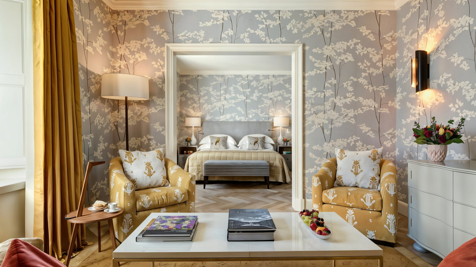Elegant sitting room with floral wallpaper, yellow armchairs, and a coffee table holding books and fruit. A cozy bed is visible in the background.