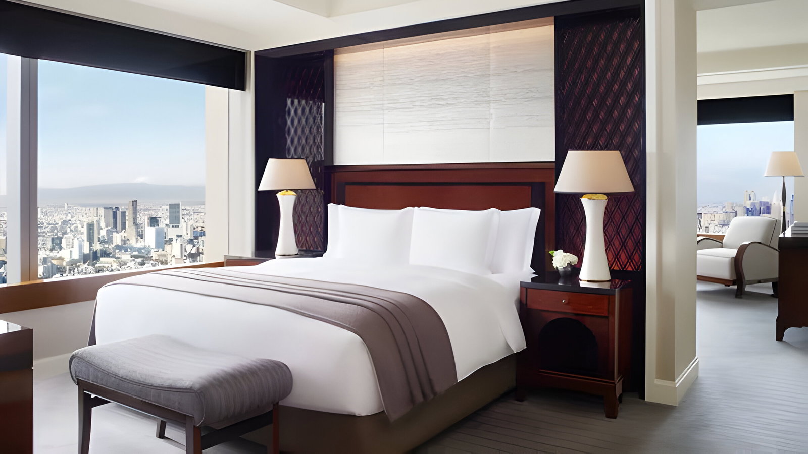 Elegant hotel room with a large bed, white linens, and brown accents. Lamps on wooden nightstands. Cityscape view through large windows.