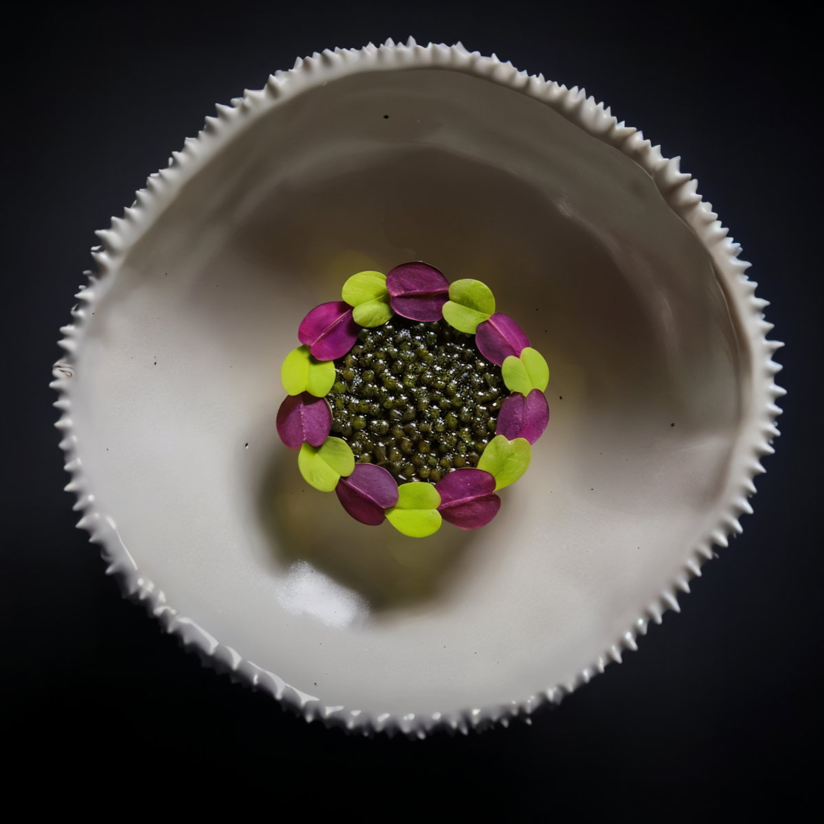 White spiked bowl with a center of green pearls, surrounded by alternating pink and green petals, on a dark background.