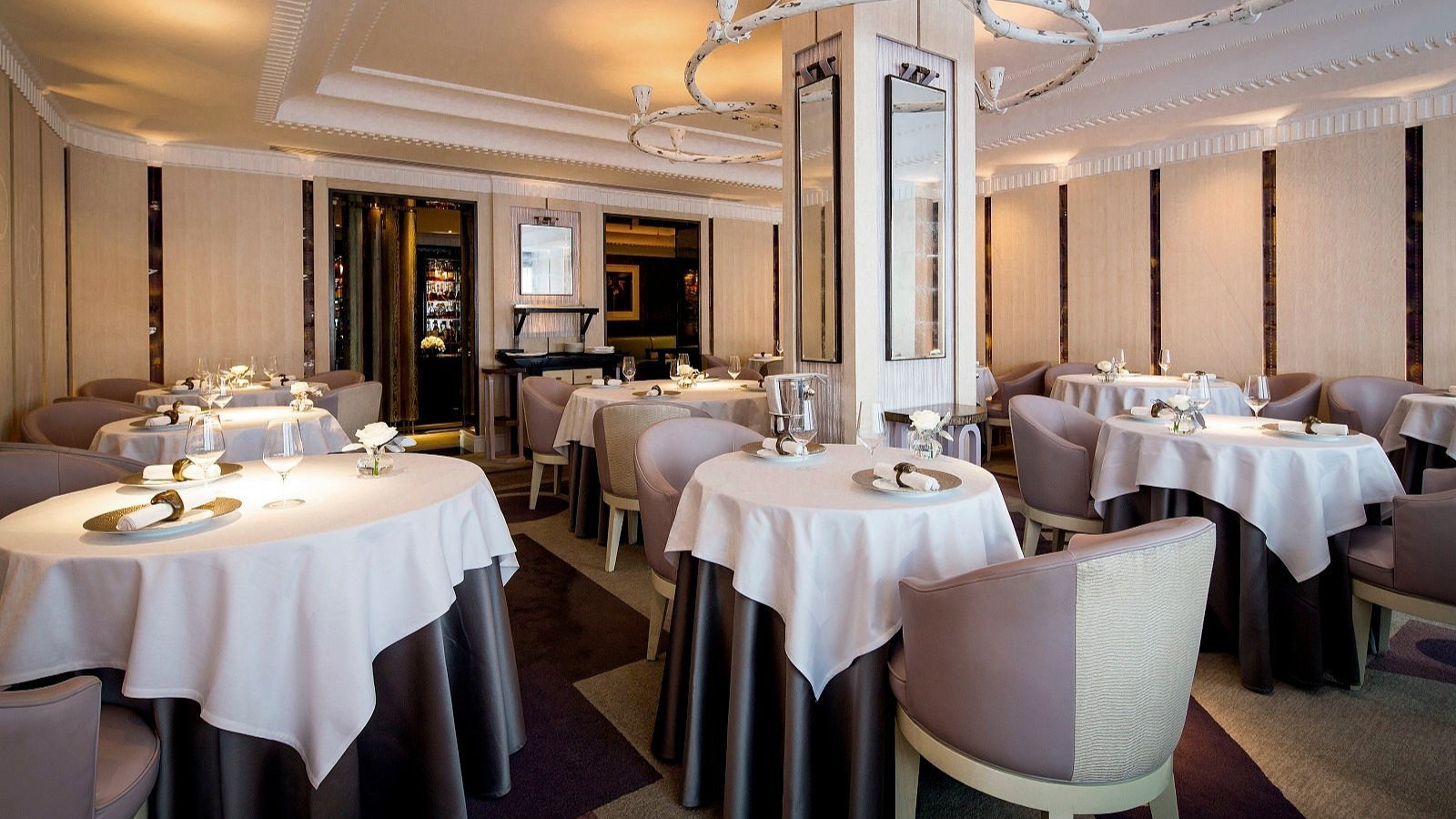 Elegant restaurant with round tables, white tablecloths, and cushioned chairs. Soft lighting and modern decor create a cozy, refined ambiance.