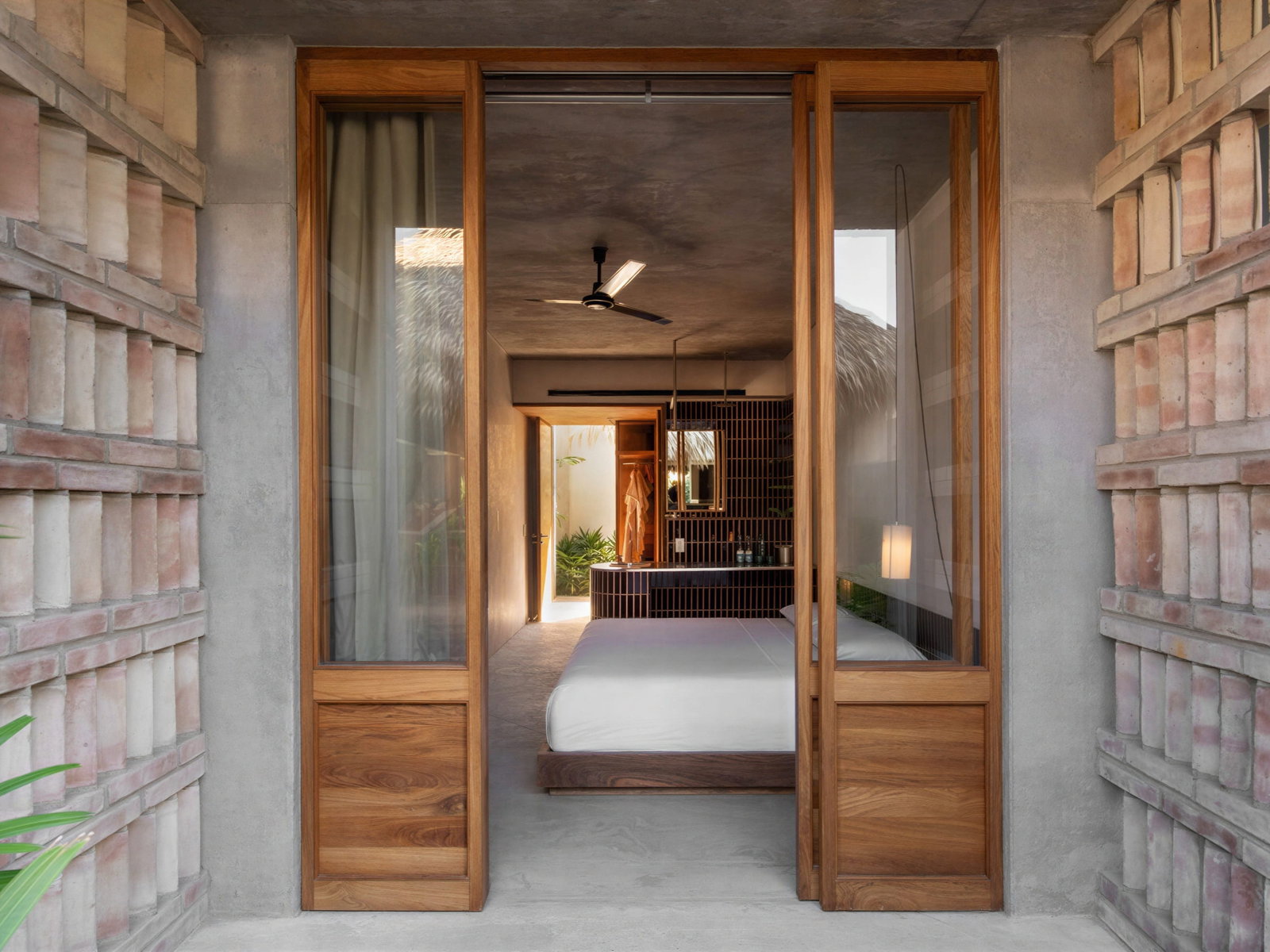 Open door view of Garden Bungalow room at Hotel Humano's minimalist design with a white bed, wooden accents, and brick walls, leading to a well-lit bathroom with greenery.