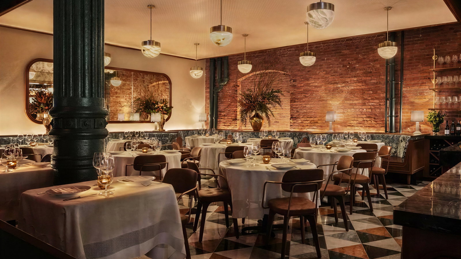 Elegant restaurant with round tables, white tablecloths, set for dinner. Dim lighting, brick walls, mirrors, and plants create a warm ambiance.